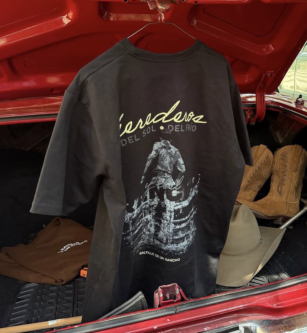 Playera negra Herederos colgada en la cajuela de un carro rojo, junto a unas botas y un sombrero
